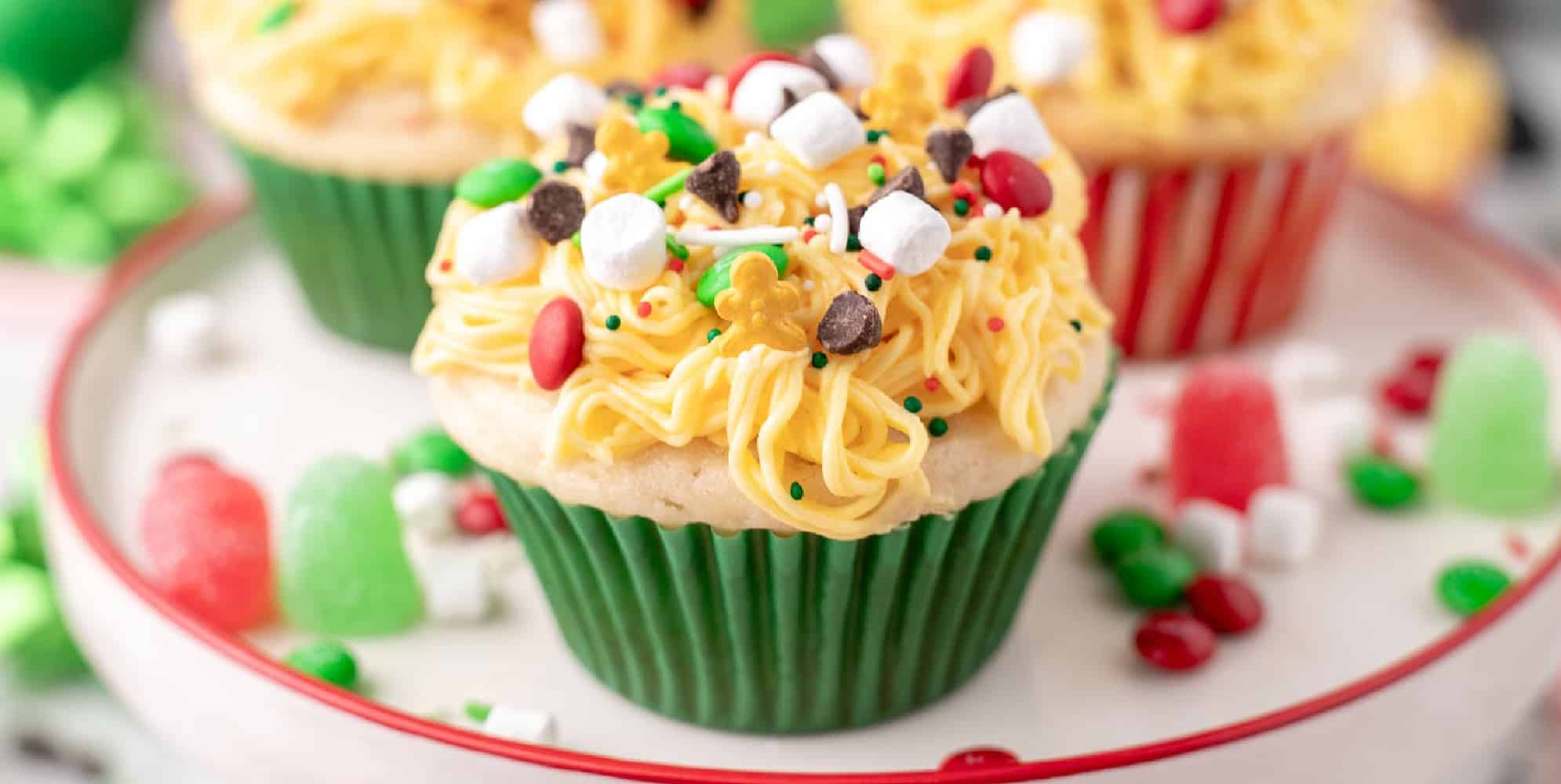 Wide view of several Elf Christmas cupcakes on a platter with more in the background, with yellow frosting made to look like spaghetti topped with candies, chocolate chips, holiday sprinkles and marshmallows.