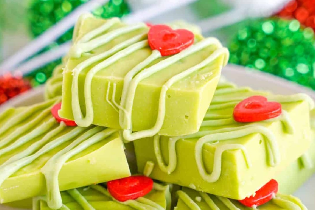 Three stacked green Grinch fudge squares with light green icing drizzles and red heart sprinkles on a plate.