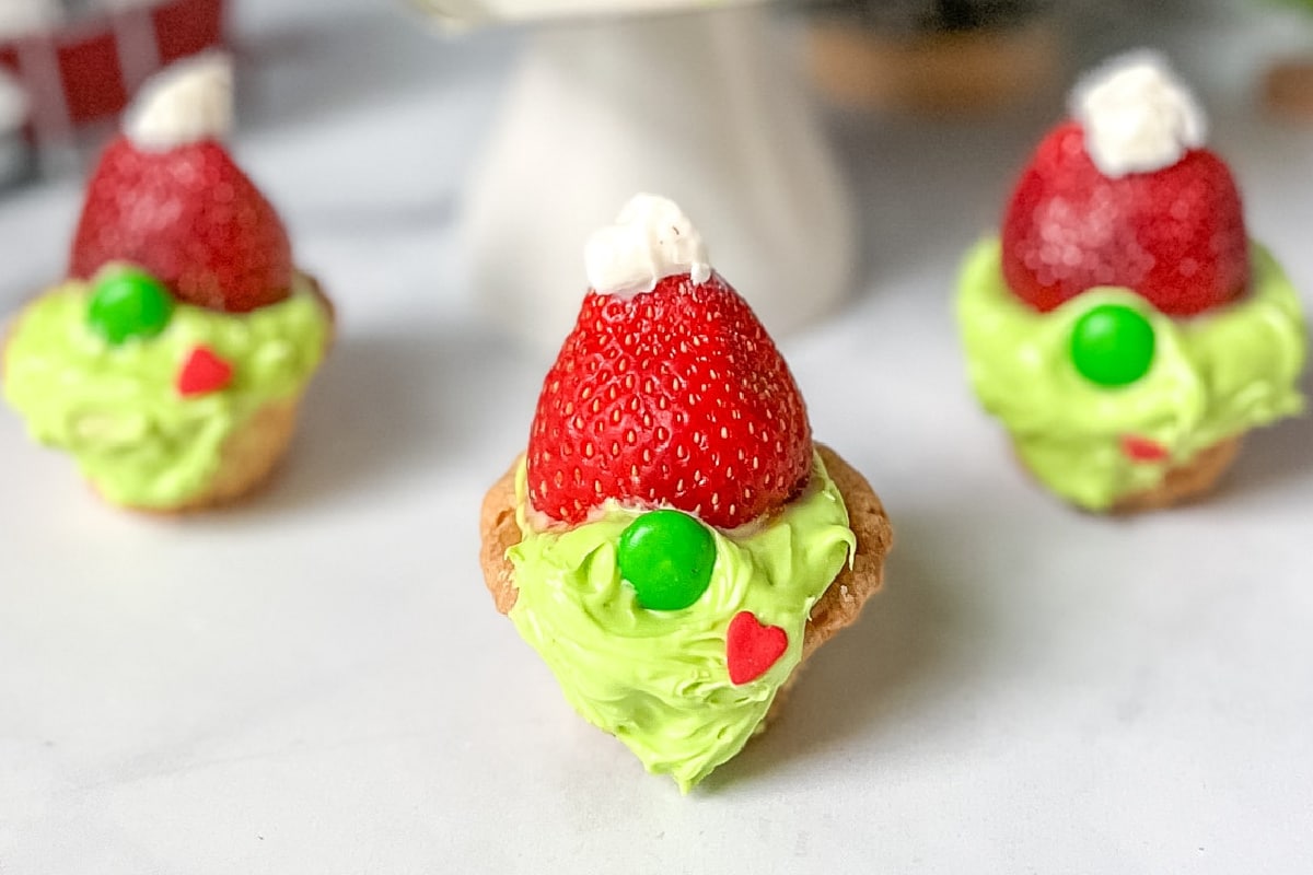 Three mini cookies topped with green frosting, strawberries, white dollops, and green and red decorations to make them look like green Grinch gnomes.