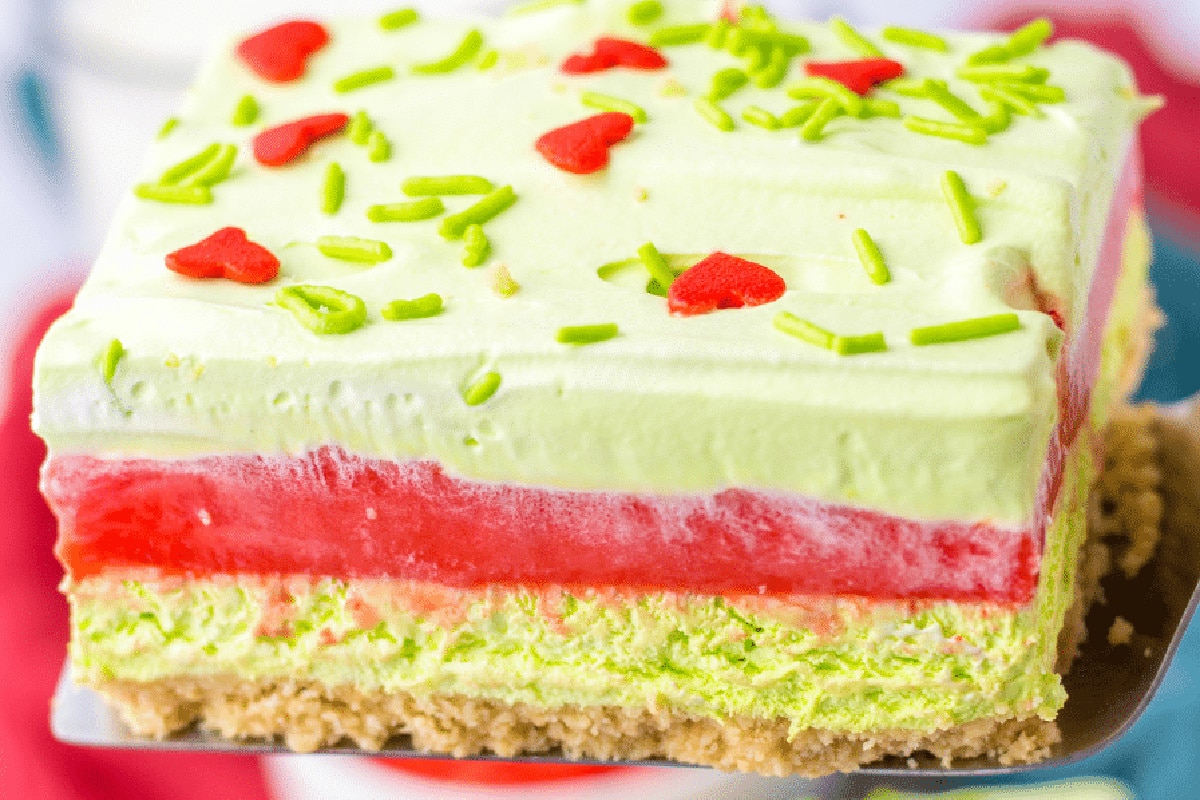 A slice of layered green and red GRinch dessert with a graham cracker crust, green and red creamy layers, topped with green sprinkles and red heart-shaped sprinkles.