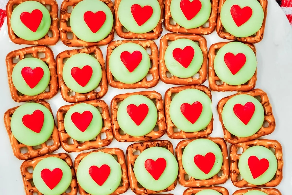 Pretzel squares topped with green candies and red heart decorations arranged in rows on a white surface.