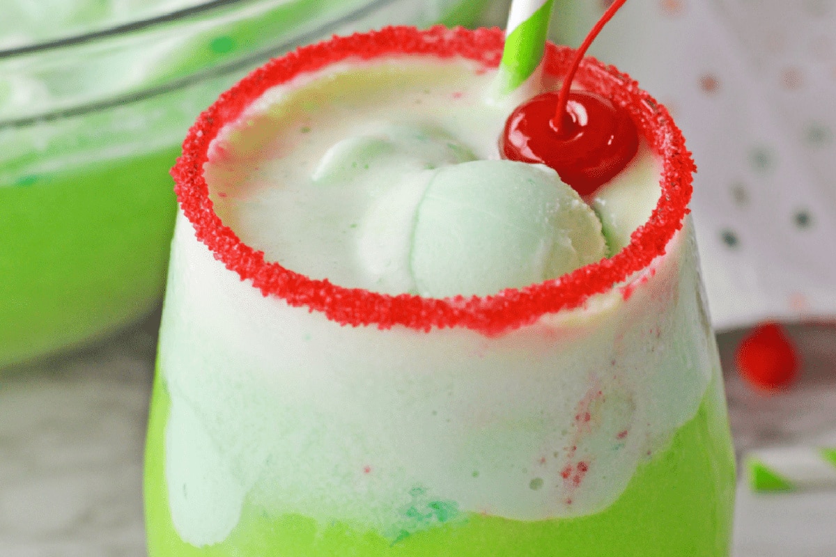 A green and white Grinch drink in a glass with a red sugared rim, topped with a red cherry and a striped straw.