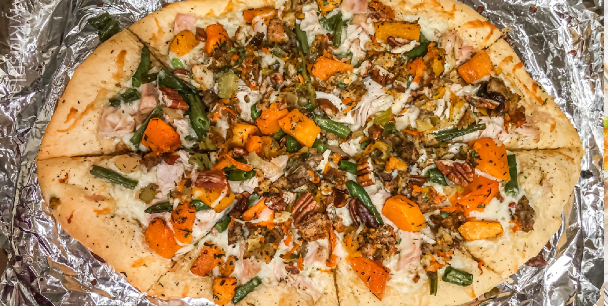 Oval pizza topped with orange squash pieces, green beans, turkey, pecans and cheese, on foil.