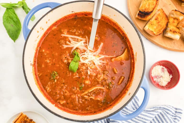 A pot of lasagna soup with a ladle on a counter from above next to a cutting board full of bread.