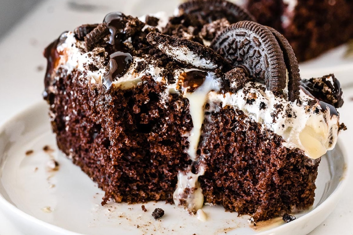 A slice of chocolate cake topped with cookies and cream frosting, drizzled with chocolate sauce, and garnished with Oreo cookie pieces on a plate.