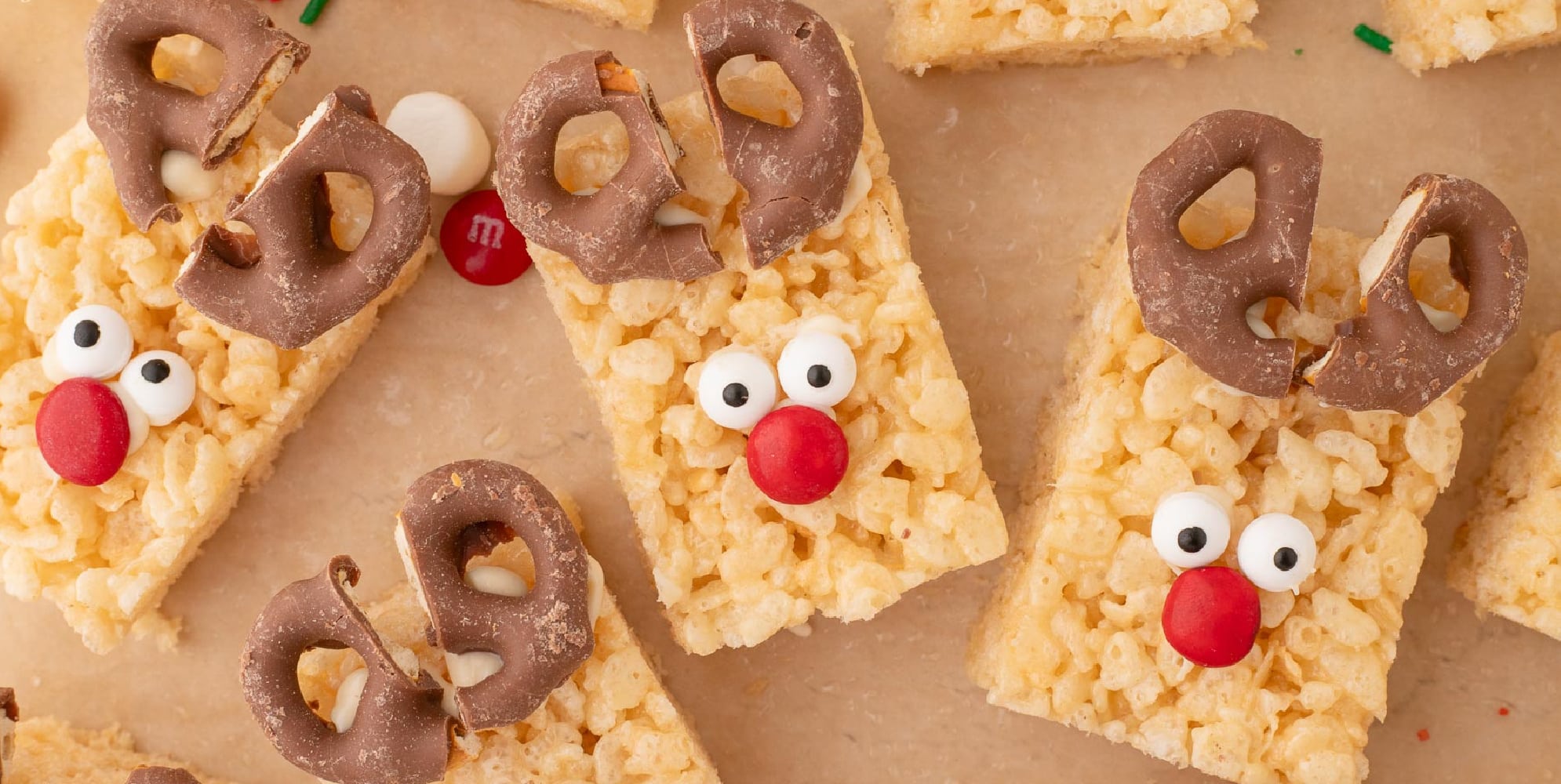 Wide view of several rectangular Rice Krispie treats decorated as reindeers with chocolate-covered pretzel antlers, candy eyes, and red candy noses.