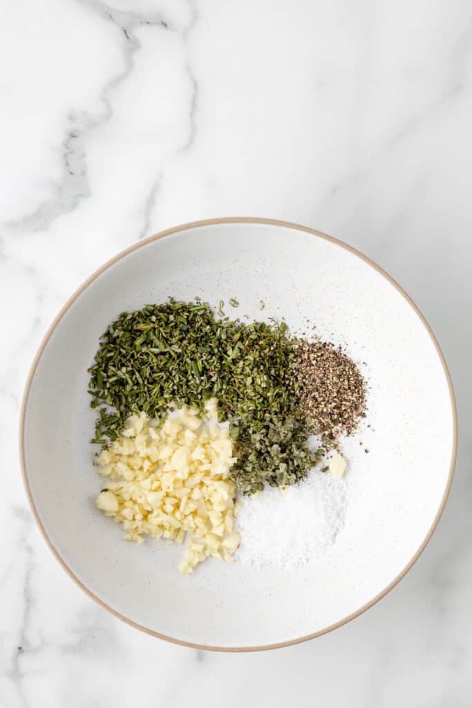 A bowl containing chopped rosemary, thyme and sage, minced garlic cloves, salt, pepper on a counter.