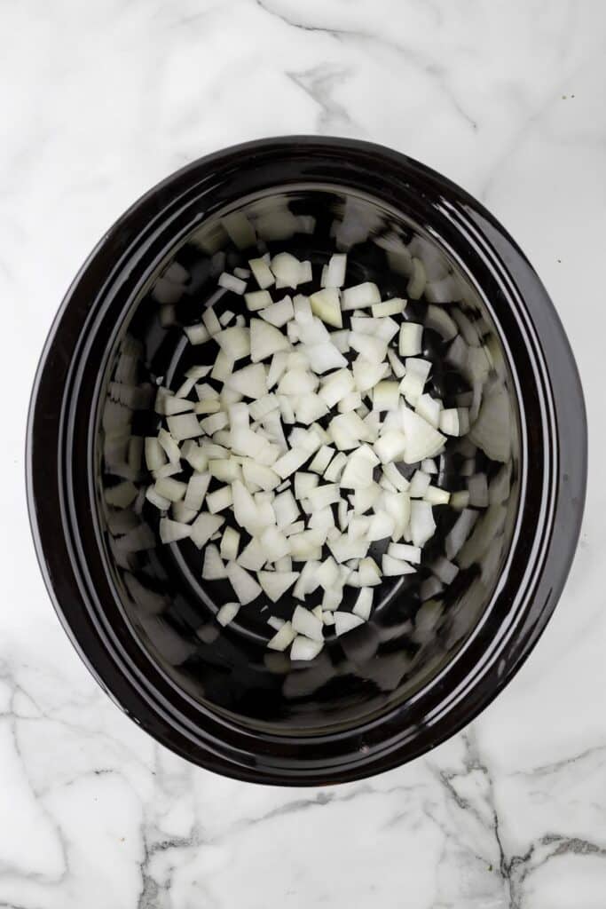 Diced onions spread out in the bottom of slow cooker on a counter from above.