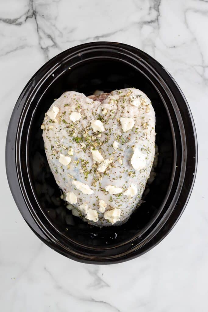 Uncooked turkey breast in a slow cooker, seasoned with herbs and topped with small butter pieces dotting the turkey.