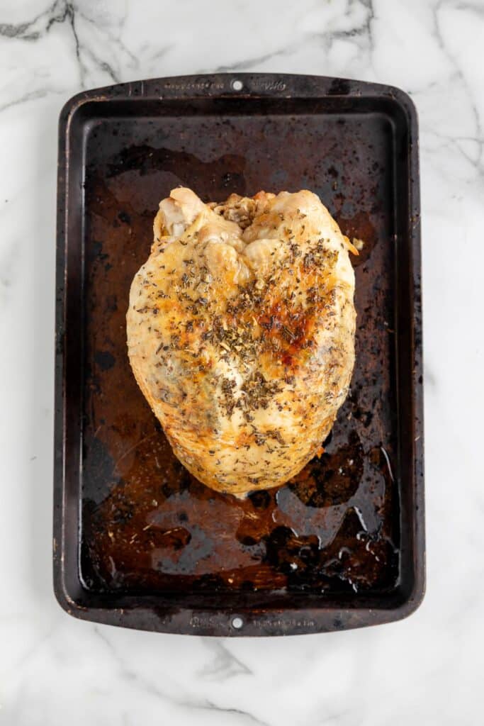 Cooked, herb seasoned turkey breast with a golden brown skin on a dark baking tray on a counter after broiling.