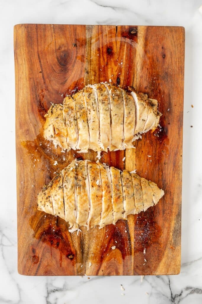 Two sliced cooked turkey breast sliced off the bone on a wooden cutting board, seasoned with herbs and spices.