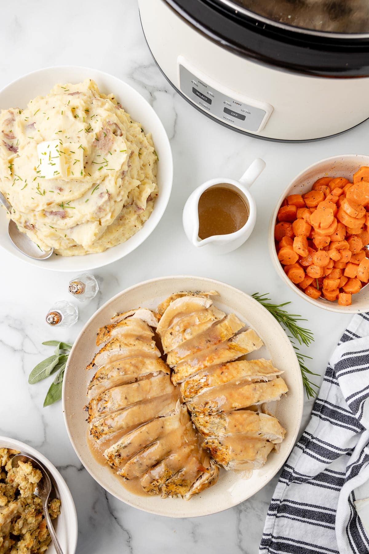 Sliced roasted turkey on a plate, surrounded by bowls of mashed potatoes, carrots, stuffing, a gravy boat, salt and pepper shakers, and a slow cooker on a table.