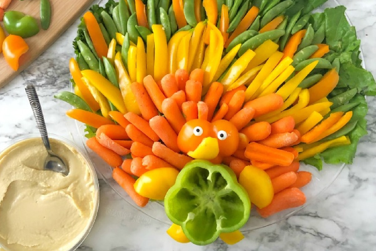 Colorful turkey-shaped veggie platter with carrots, bell peppers, and snap peas arranged in fan shape. Hummus dip in a bowl on the side.