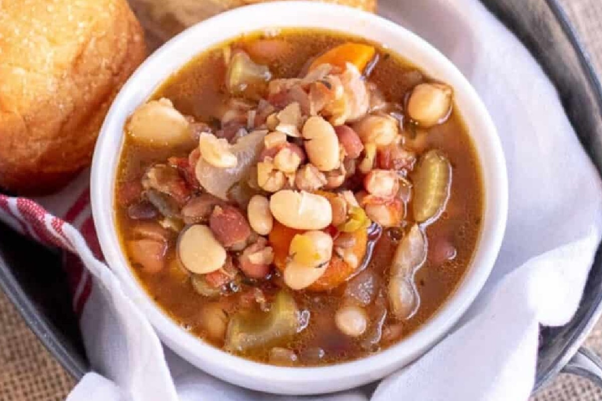 A bowl of mixed bean soup with vegetables, such as carrots and celery, garnished with a slice of bread on the side.