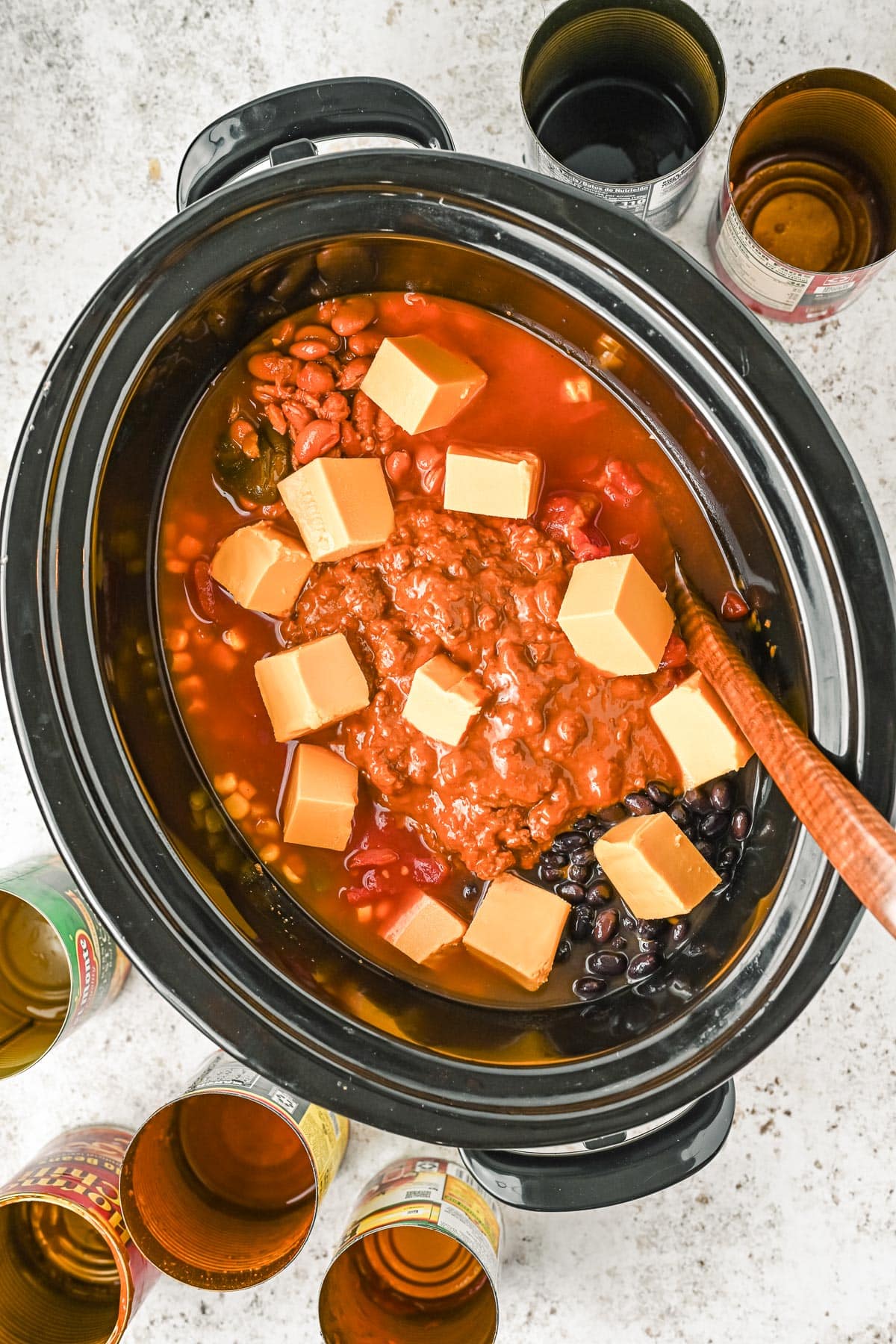 Slow cooker full of beans, tomatoes, Velveeta cheese and other ingredients for 7 can chili with open cans nearby.