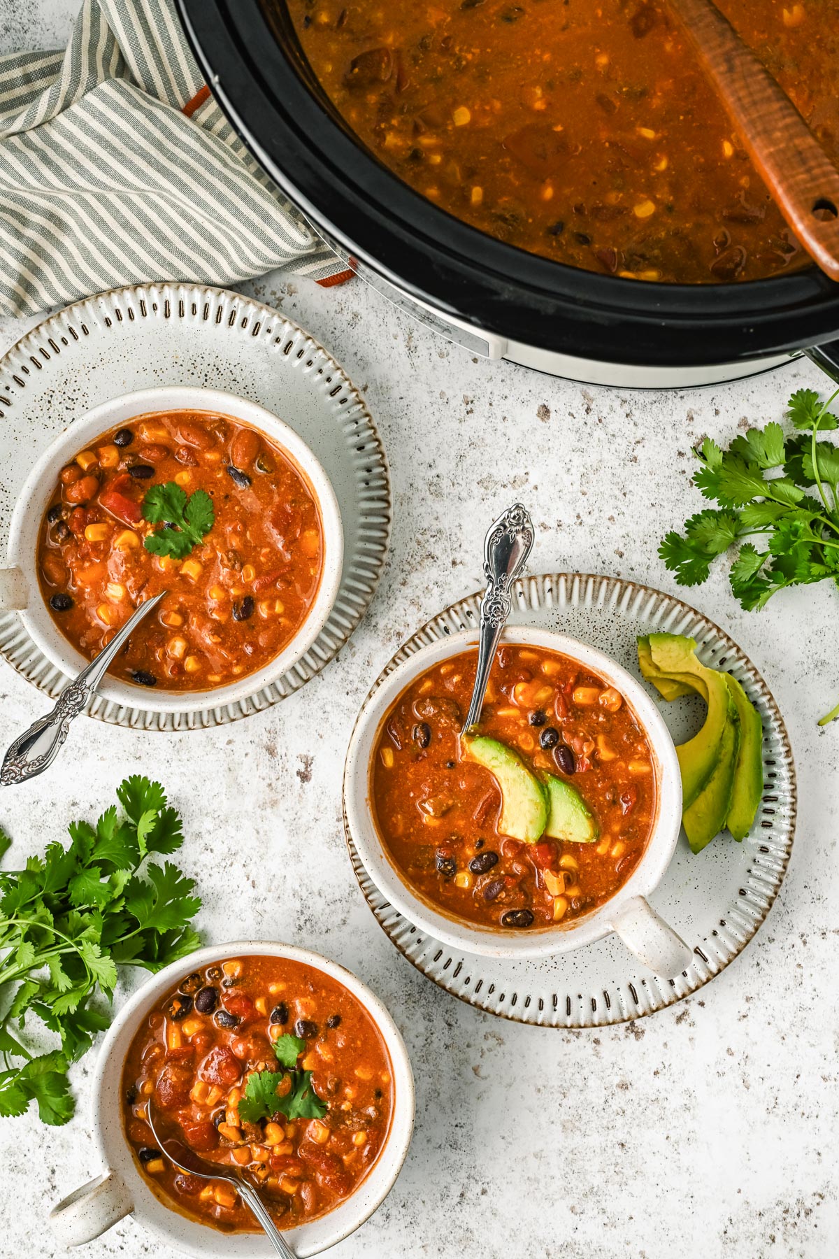 Three bowls of 7 can soup with beans and corn are on a table next to slices of avocado and cilantro, with a slow cooker full of more 7 can soup nearby on the counter.