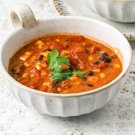 A square view of a bowl filled with chili soup containing corn, beans, and chunks of tomato, garnished with cilantro.