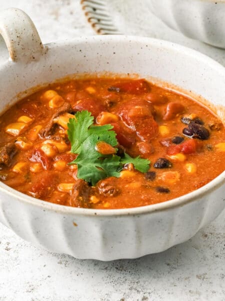 A square view of a bowl filled with chili soup containing corn, beans, and chunks of tomato, garnished with cilantro.