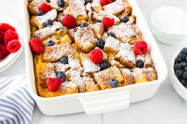 Baked French toast casserole topped with powdered sugar, raspberries, and blueberries in a white dish.