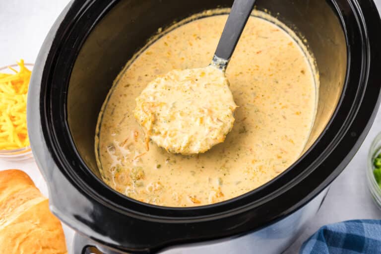 Wide view of slow cooker with creamy soup ladled up, surrounded by cheddar cheese, bread, and a napkin near the crock pot.