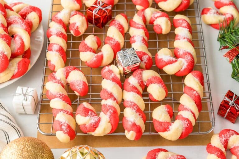 Red and white twisted cookies shaped like candy canes on a cooling rack, surrounded by decorative gift boxes and ornaments.