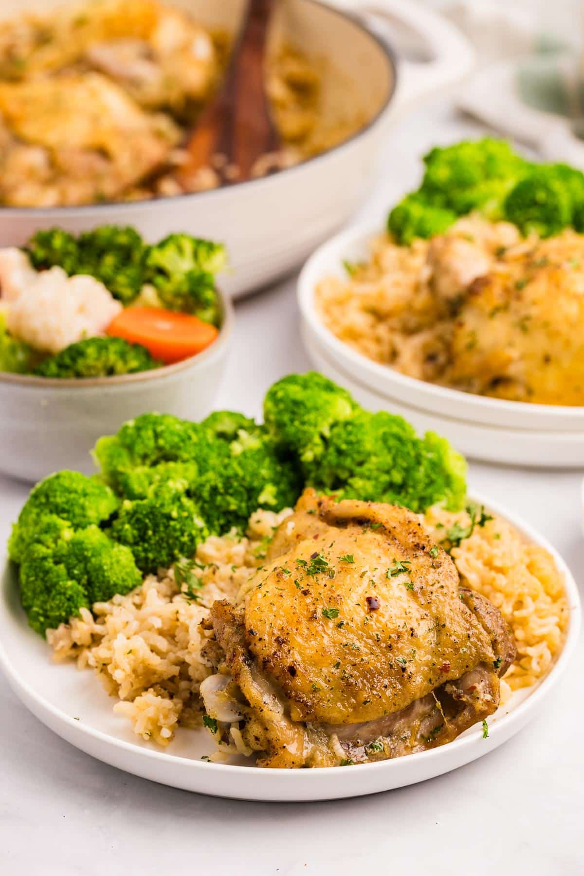 Plate of seasoned chicken thighs on rice with a side of broccoli, and more vegetables and the pan of chicken and rice in the background on the table.