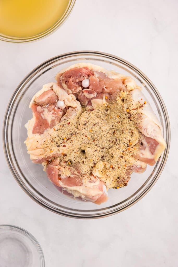 A glass bowl with raw chicken thighs being mixed with spices for chicken thighs and rice on a counter.