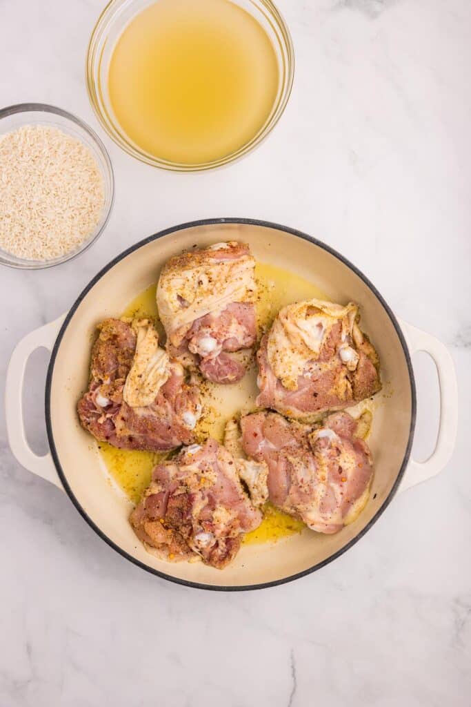 Raw seasoned chicken thighs skin side down in a pot with oil next to a bowl of rice and a bowl of chicken broth on a counter.