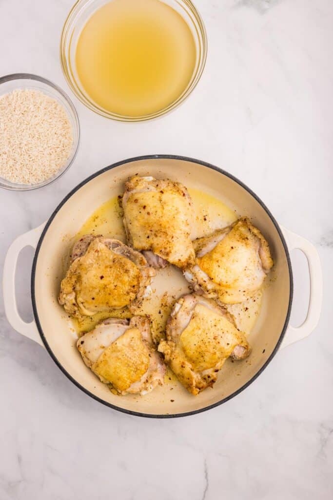 Five seasoned raw but browned chicken thighs in a white pot, beside uncooked rice and a bowl of broth on a counter