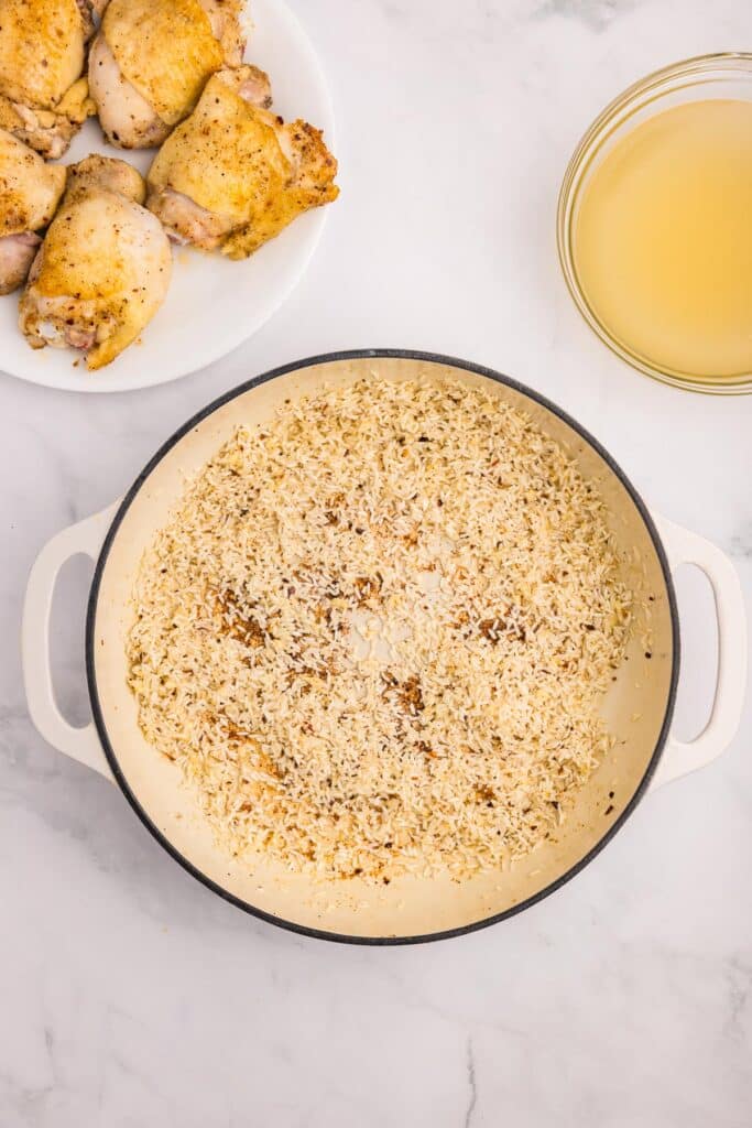 A pot with sautéed rice and garlic with a plate of browned bone in chicken thighs and a bowl of chicken broth nearby for chicken thighs and rice.