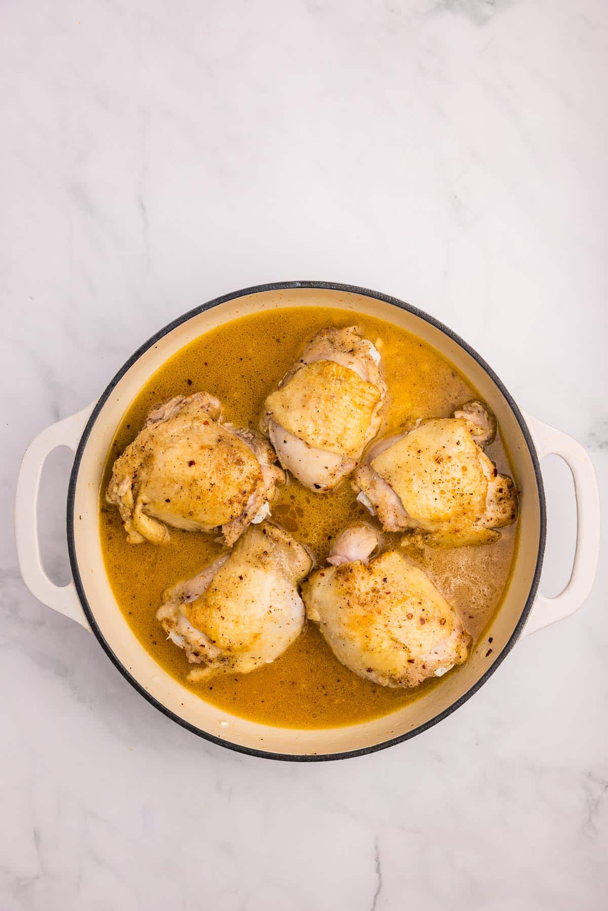 A white pot containing browned chicken thighs sitting in a pot full of broth and rice before baking.