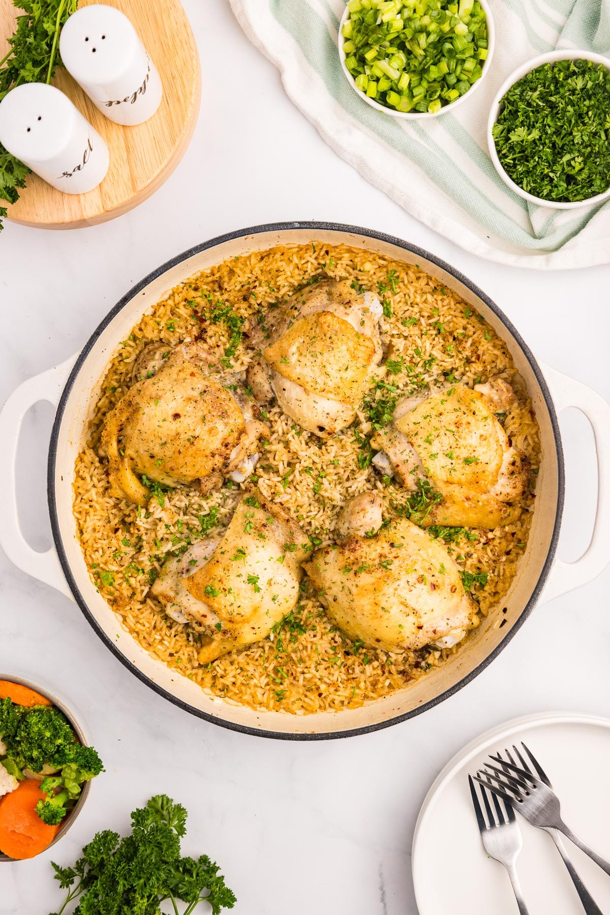 A skillet with cooked chicken thighs and rice topped with seasoning with plates, forks and other items for dinner on the counter nearby.