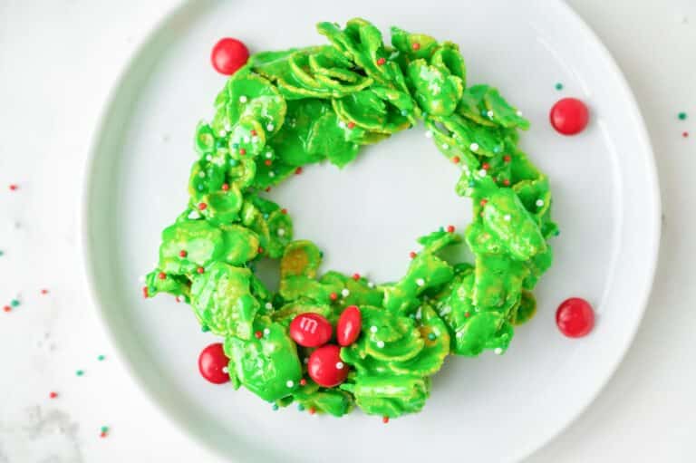 A wide view of a green wreath-shaped cereal treat made of cornflakes decorated with red candies and sprinkles on a plate.