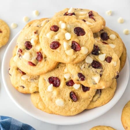 Wide view of a plate of cookies with white chocolate chips and dried cranberries, surrounded by more cookies and a small bowl of cranberries.