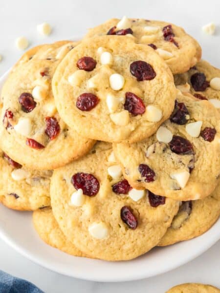 Wide view of a plate of cookies with white chocolate chips and dried cranberries, surrounded by more cookies and a small bowl of cranberries.