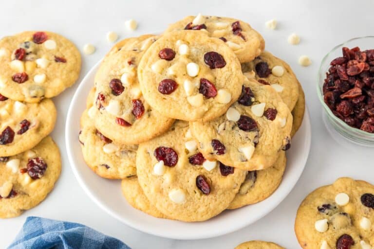 Wide view of a plate of cookies with white chocolate chips and dried cranberries, surrounded by more cookies and a small bowl of cranberries.