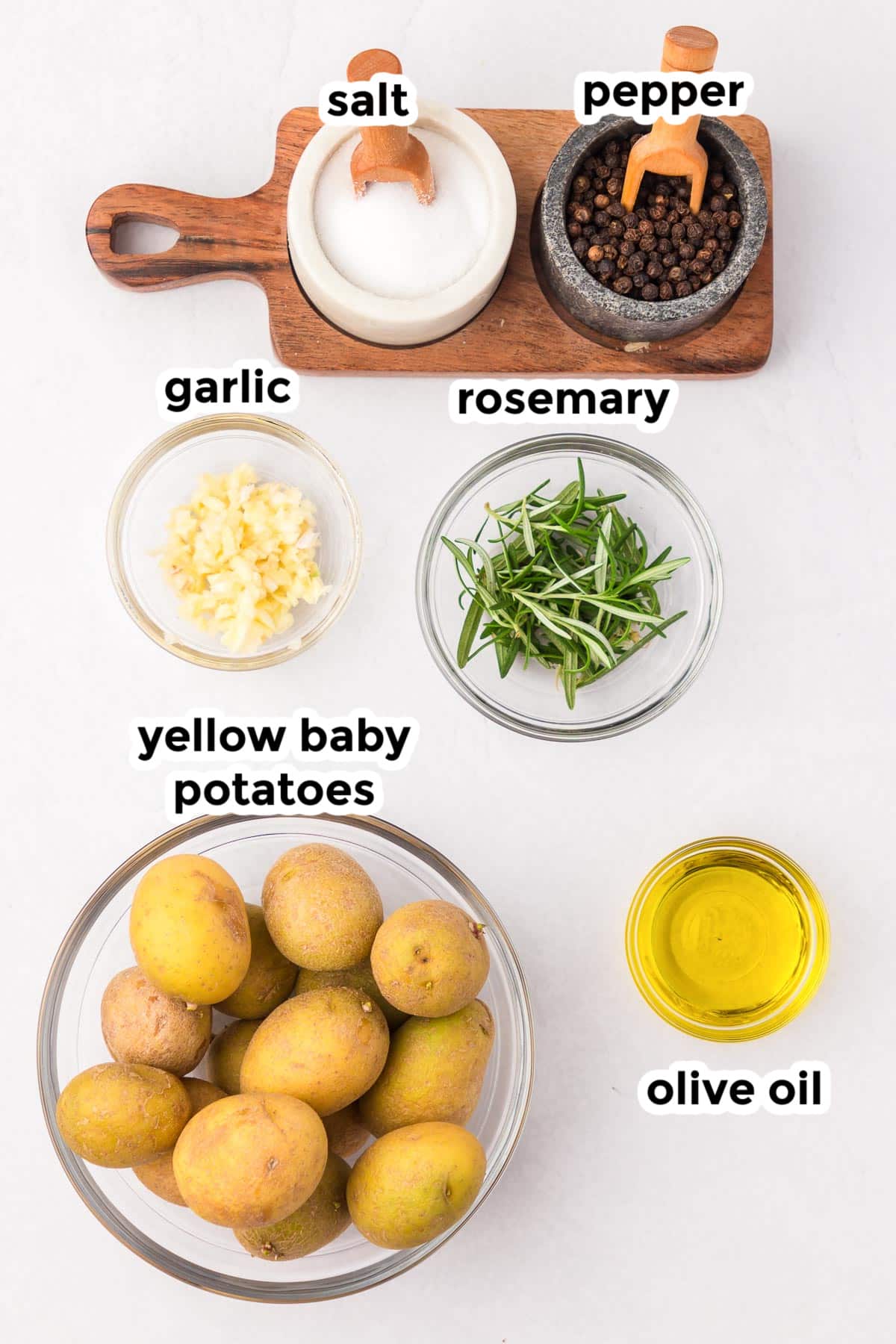 Ingredients laid out: salt, pepper, minced garlic, rosemary, yellow baby potatoes in bowls, and olive oil on a white surface.