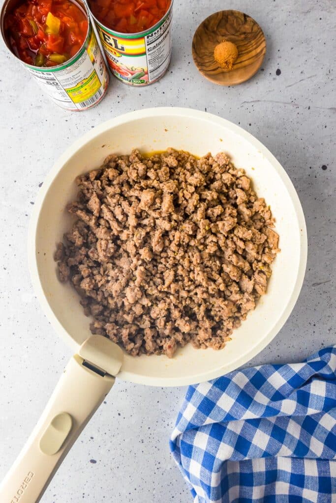 Cooked ground sausage in a skillet with open cans of Rotel tomatoes with green chilis and taco seasoning on the counter nearby for crock pot rotel dip.