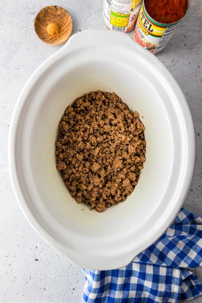 A slow cooker filled with cooked pork with open cans of Rotel and taco seasoning on the counter nearby for crock pot Rotel dip.