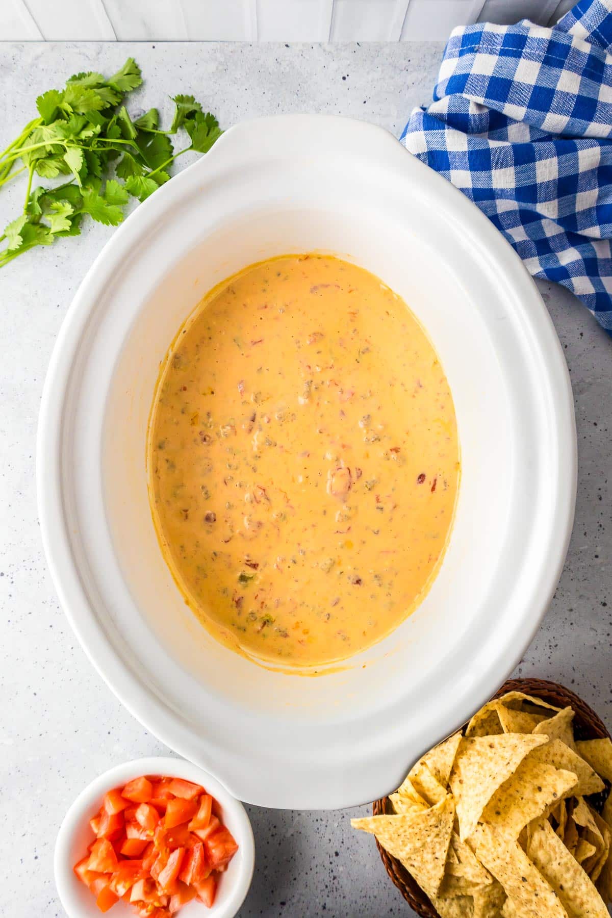 A crockpot filled with creamy Rotel dip on a countertop with tortilla chips and diced tomatoes in bowls nearby.