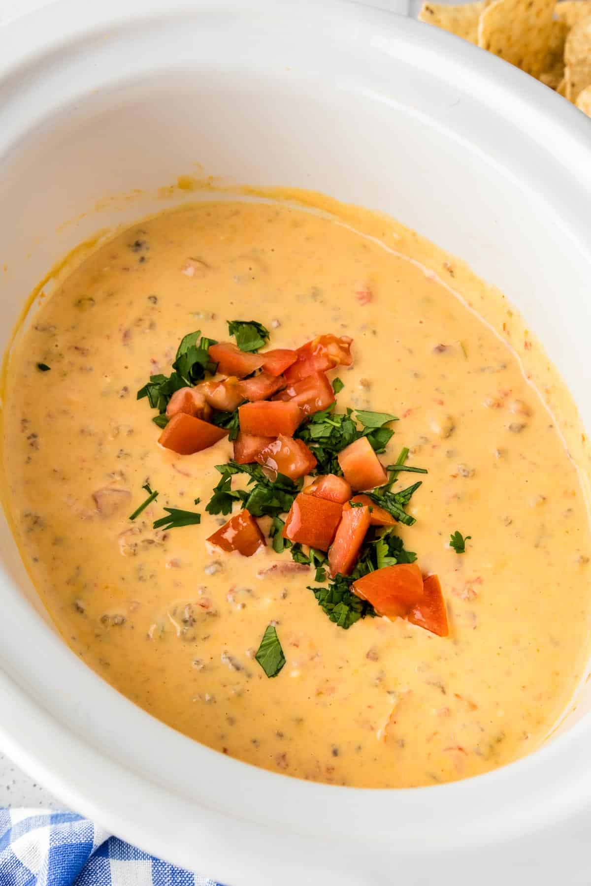 A white crock pot filled with creamy cheese rotel dip, topped with chopped tomatoes and cilantro.