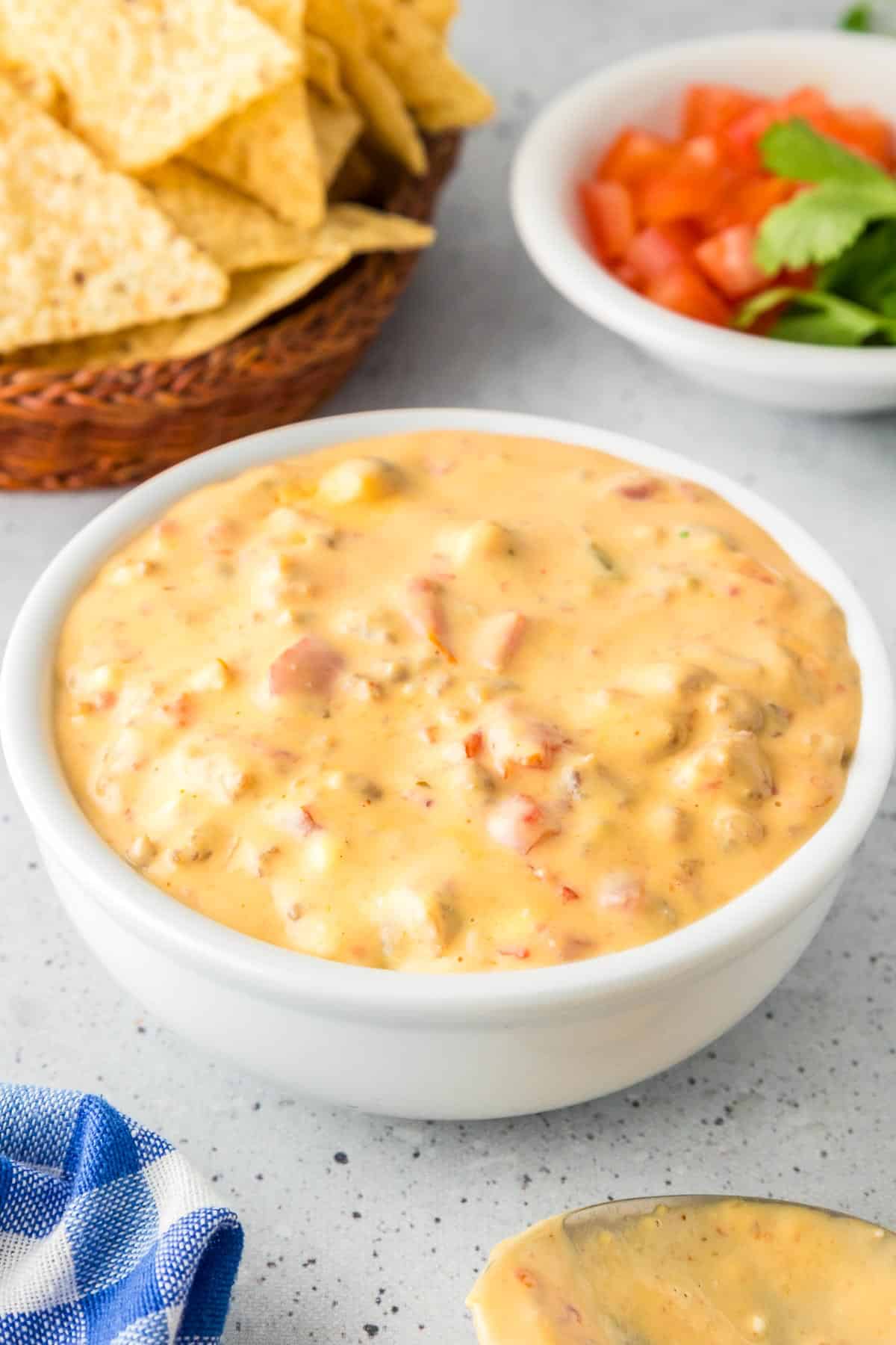 A bowl of creamy cheese crock pot rotel dip with visible chunks of sausage, tomatoes and peppers with tortilla chips and diced tomatoes nearby on the counter in bowls.