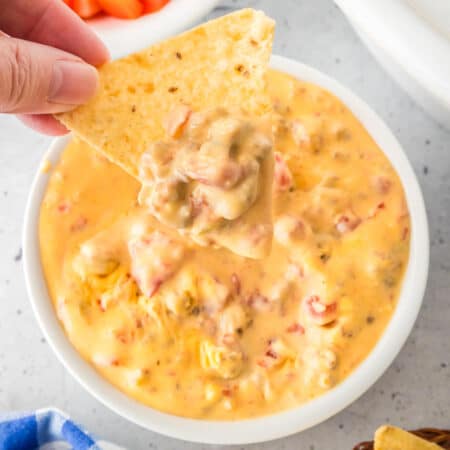 A hand holds a tortilla chip dipped in a bowl of creamy rotel dip next to a slow cooker on a counter.