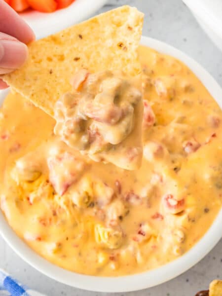 A hand holds a tortilla chip dipped in a bowl of creamy Rotel dip next to a slow cooker on a counter.