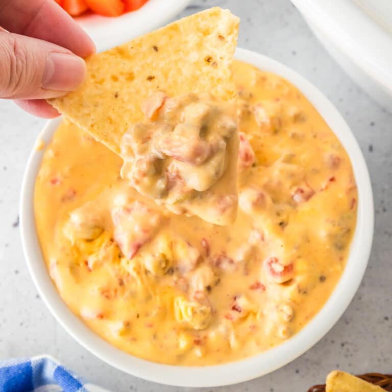 A hand holds a tortilla chip dipped in a bowl of creamy Rotel dip next to a slow cooker on a counter.