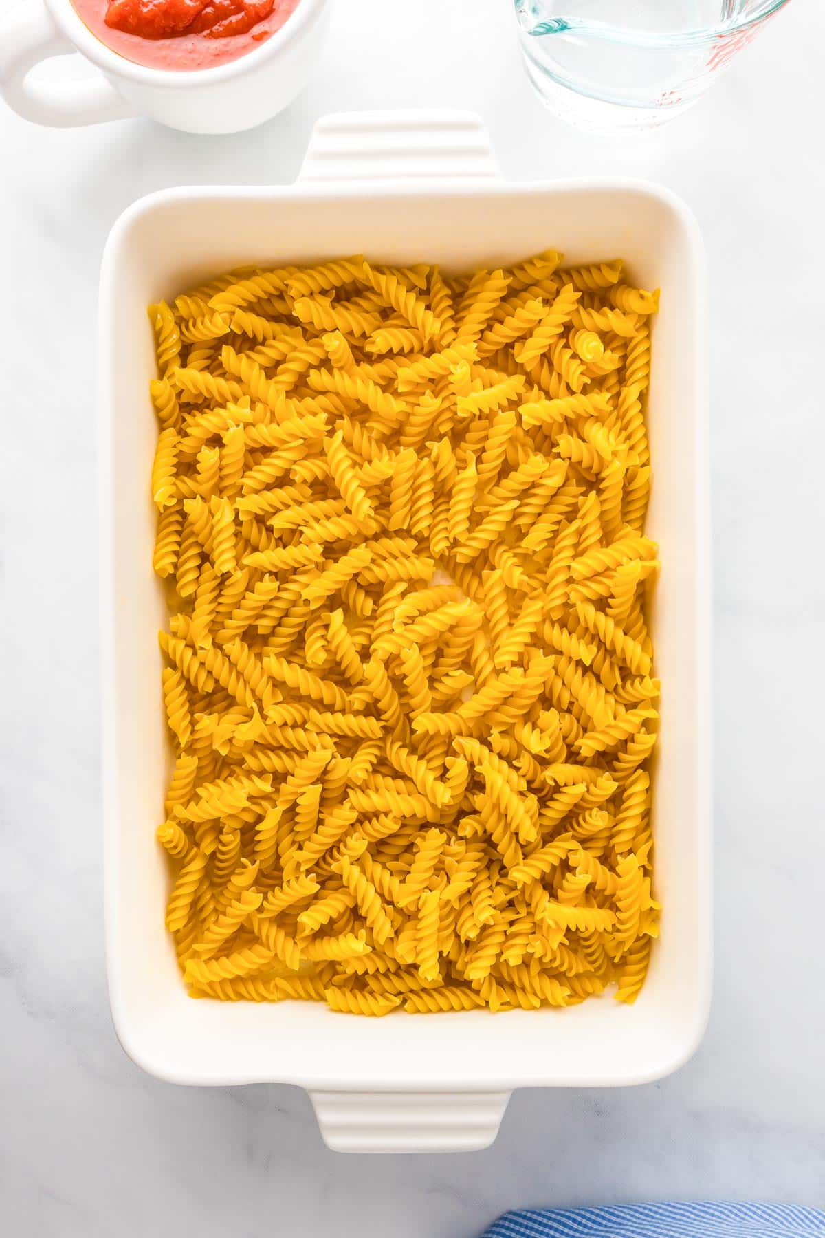 Uncooked fusilli pasta in a white rectangular baking dish on a countertop, with a small bowl of red sauce in the corner.