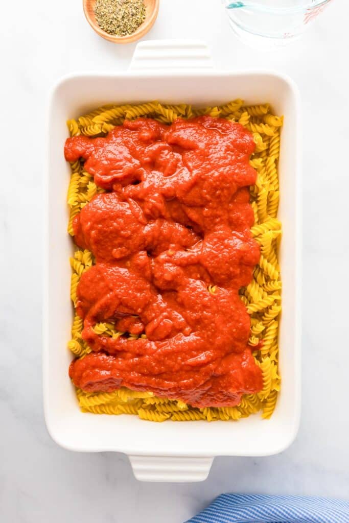 A white baking dish with uncooked fusilli pasta topped with red tomato sauce. A small bowl of herbs and a glass of water are nearby.