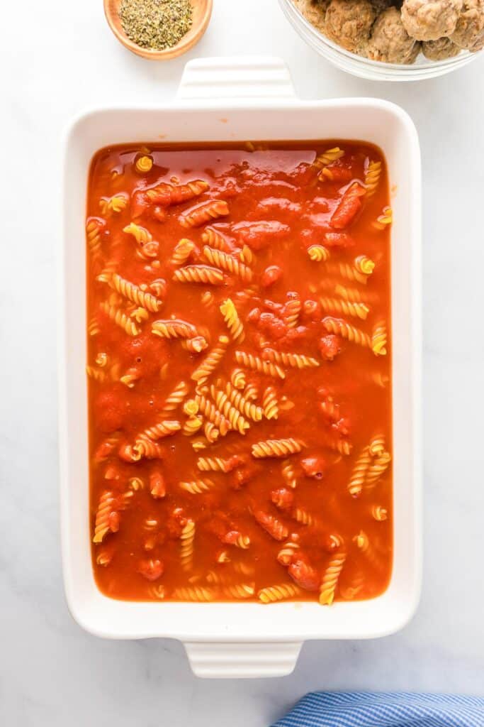 Baking dish with uncooked rotini pasta in tomato sauce, on a white surface near a bowl of meatballs and a small dish of herbs.