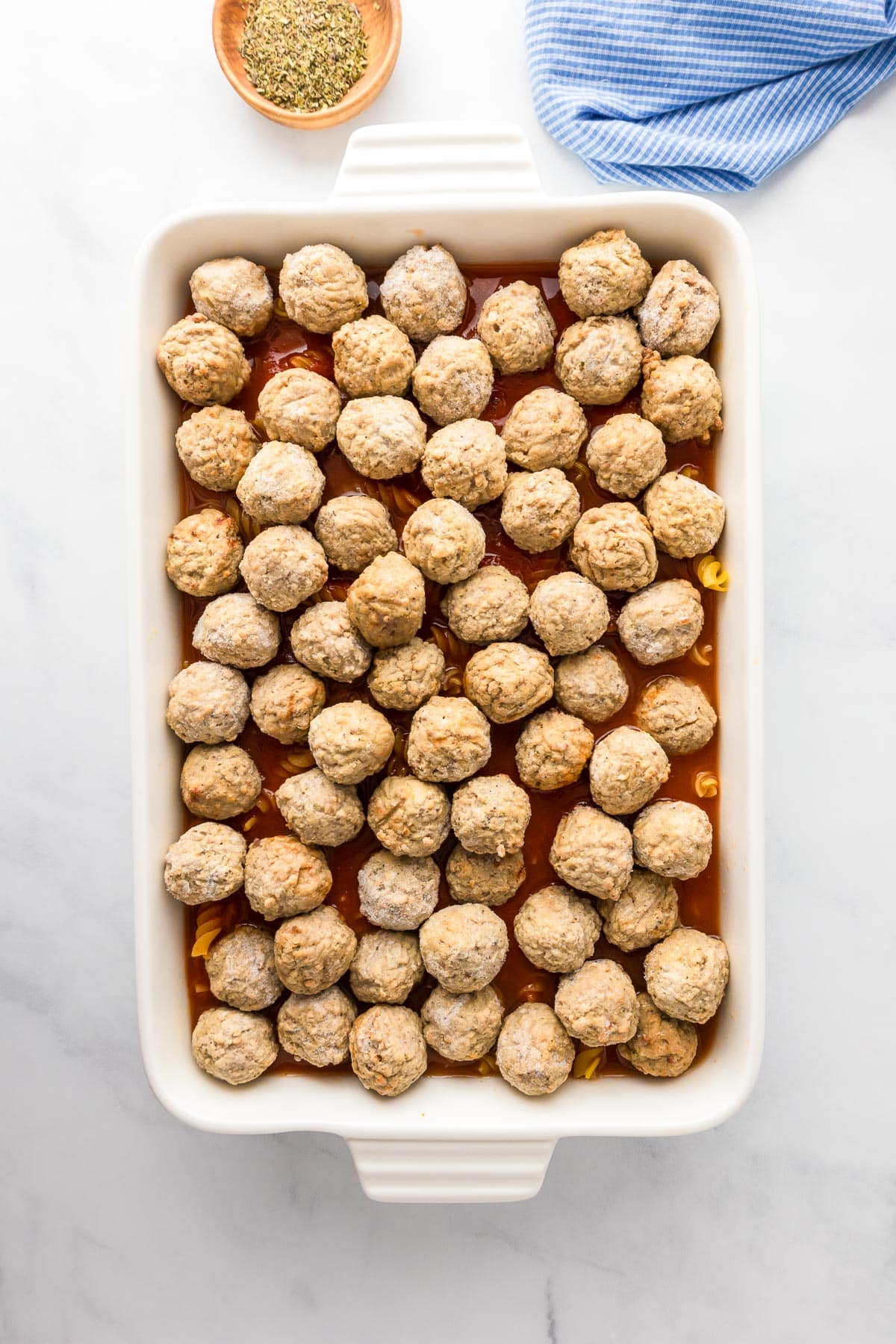 A baking dish filled with evenly arranged meatballs on a layer of sauce, with a small bowl of herbs and a blue cloth nearby.