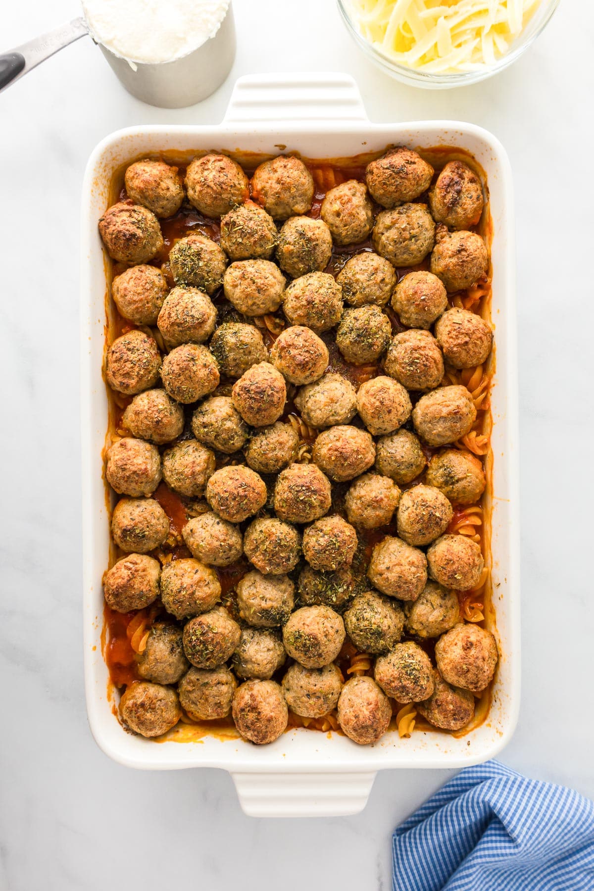 A white baking dish filled with evenly spaced meatballs over layers of pasta and sauce. Grated cheese and a cup of flour are visible in the background.